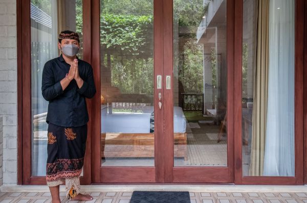Balinese man in traditional batik sarong and face mask greets with prayer hands at open wooden villa doors, revealing tropical bedroom interior.